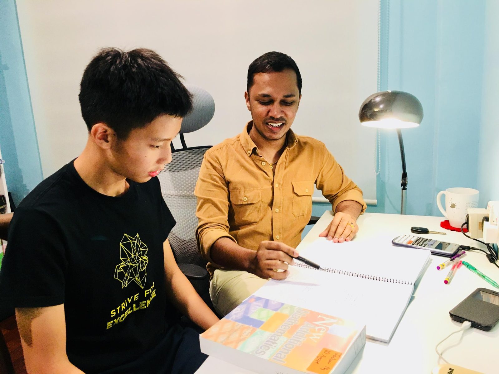 Mr Kumaraveelu guiding a student through a Mathematics workbook at a study desk